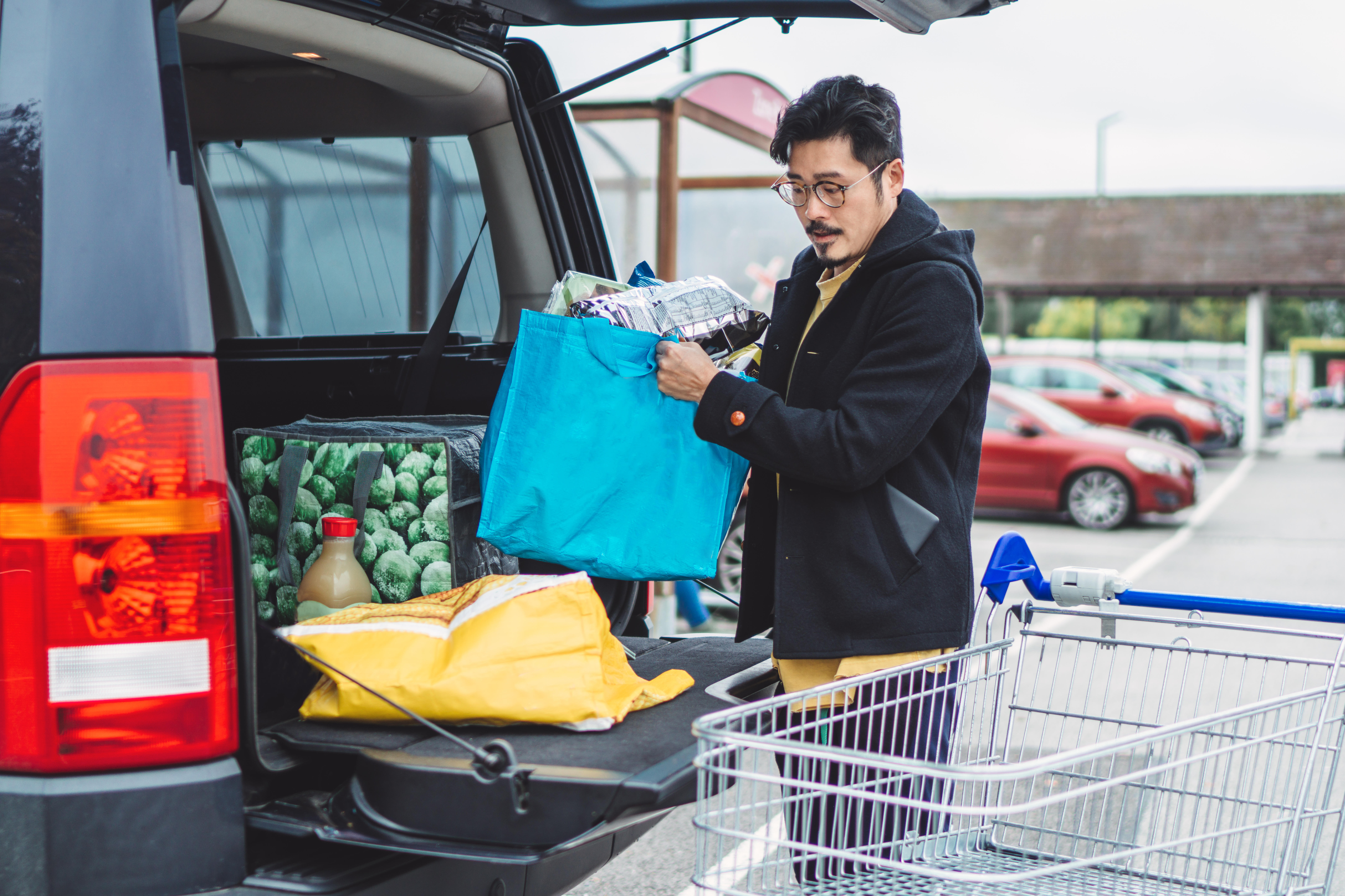 Young man putting away bags of groceries in his car trunk at supermarket car park 