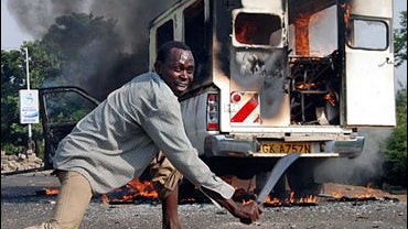 An opposition supporter scrapes a machete along the road next to a burning vehicle 