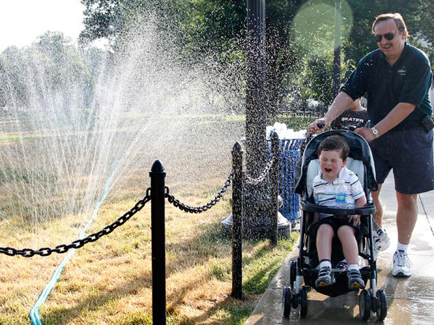 pushes his son on a stroller through a water sprinkler to cool off 
