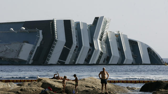 Tourists relax on rocks in front of the Costa Concordia wreckage  