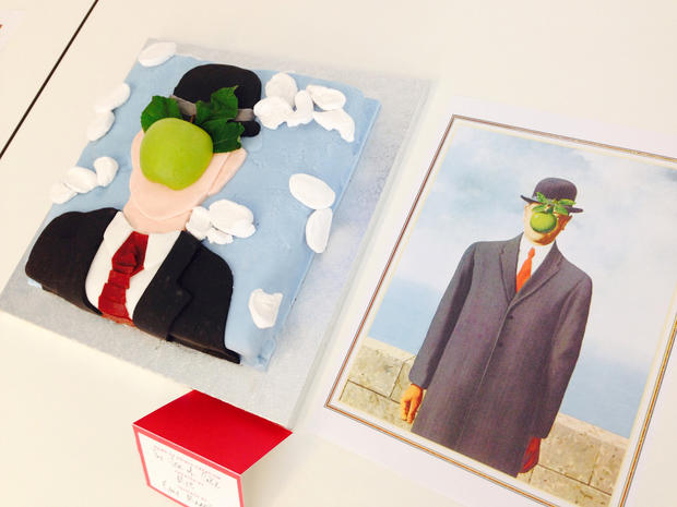 A cake inspired by Rene Magritte's "The Son of Man" is seen next to a picture of the painting. 