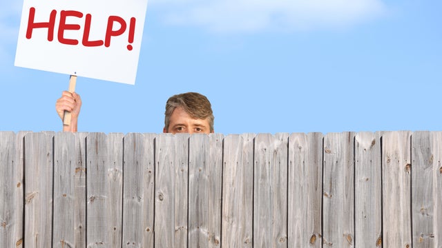 Man with HELP sign looking over wood privacy fence 