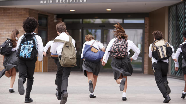 Group Of High School Students Wearing Uniform Running Into School Building At Beginning Of Class 