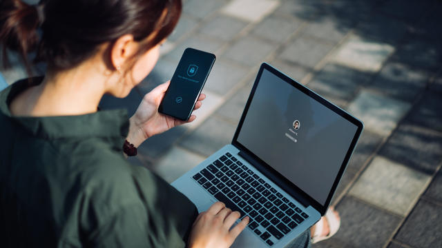 Young Asian businesswoman sitting on the bench in an urban park working outdoors, logging in to her laptop and holding smartphone on hand with a security key lock icon on the screen. Privacy protection, internet and mobile security concept 