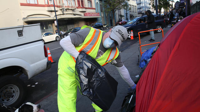 John Duport, Department of Public Works worker, hands a trash bag to someone in a tent on Hyde Street while cleaning the street and sidewalk on Wednesday, March 25, 2020 in San Francisco, Calif. 