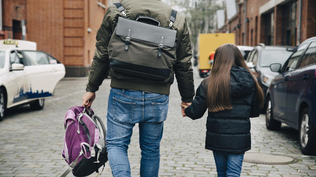 Rear view of father and daughter with backpack walking on footpath in city 