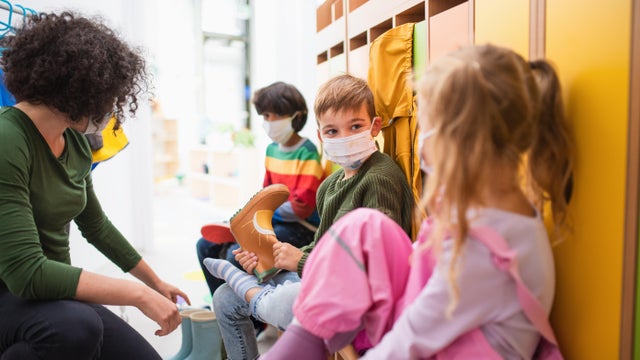 Pre school teacher helping children to put on shoes indoors in cloakroom at nursery, coronavirus concept. 