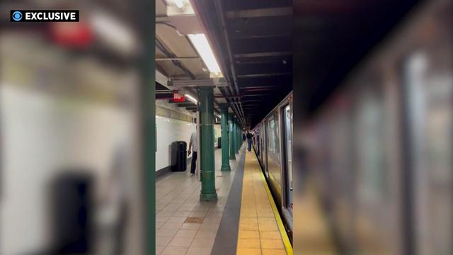 A man on a New York City subway platform holds a gun in his left hand while arguing with another man on an empty subway train. 