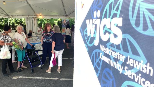 The foreground of the photo shows a sign that reads "WJCS: Westchester Jewish Community Services." In the background are several set tables with several people standing and sitting around, socializing. 