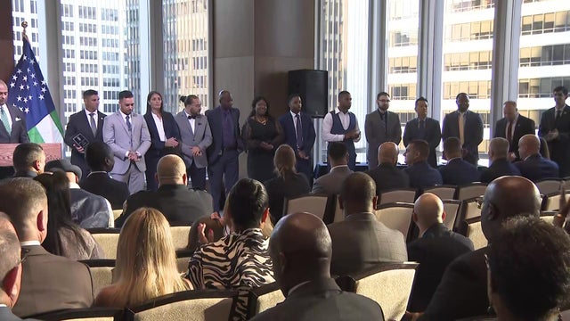 About a dozen members of the NYPD, all wearing formal attire, stand in a row on a stage in front of an audience. 