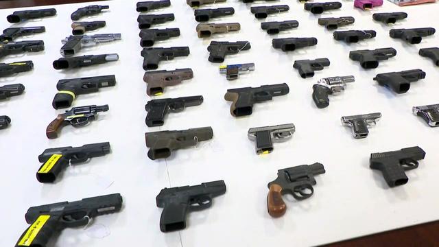 Dozens of firearms are displayed on a white table. 