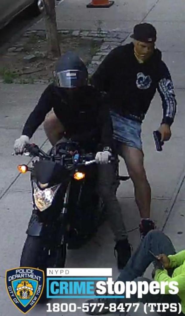 A still from surveillance video shows two individuals on a motorcycle. The passenger is pointing a firearm at an individual sitting on the sidewalk. 