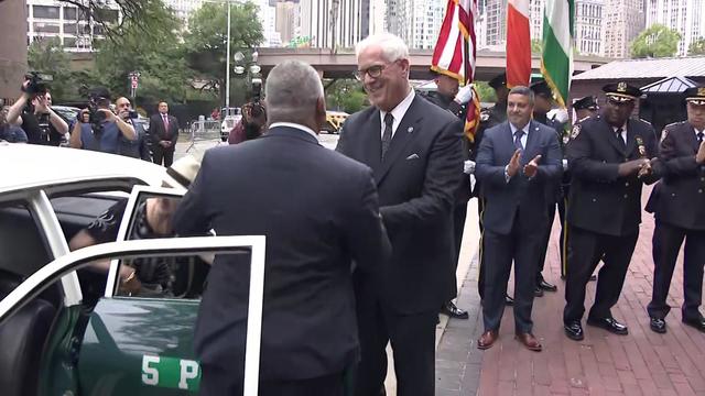 John Miller shakes hands with an NYPD official as other officials and officers stand behind them, clapping. 