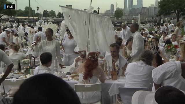 Dozens of people dressed in all white sit at tables in Battery Park for Dîner en Blanc. 
