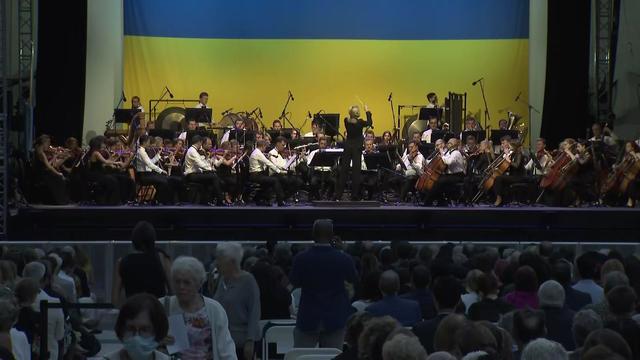 An orchestra performs on an outdoor stage in front of a large Ukrainian flag. 