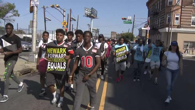 A crowd walks through the streets of Newark, some holding signs reading, "Newark forward minus violence equals our future." 