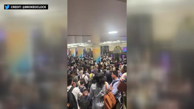 Dozens of people stand packed shoulder-to-shoulder in Penn Station. 