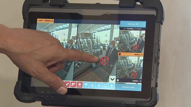 A screen on a tablet shows footage from a security system indicating someone walking between metal detectors has a weapon. 