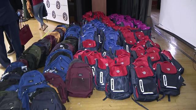 Dozens of backpacks sit on the floor of a school auditorium's stage. 