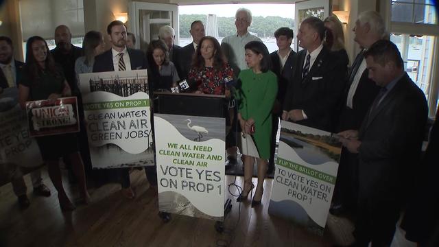 A group of lawmakers and advocates stand around a podium with signs that say "Vote yes on Prop 1. Clean water, clean air, green jobs." 
