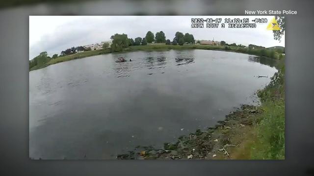 A screenshot from body camera footage shows a vehicle partially submerged in a pond. 