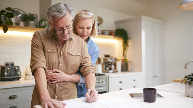 Mature Couple Reviewing And Signing Domestic Finances And Investment Paperwork In Kitchen At Home 