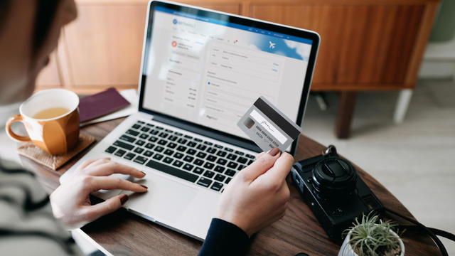 Over the shoulder view of young Asian woman shopping online for flight tickets on airline website with laptop, entering credit card details to make mobile payment at home. Camera and passport on the table. Travel planning. Booking a holiday online 