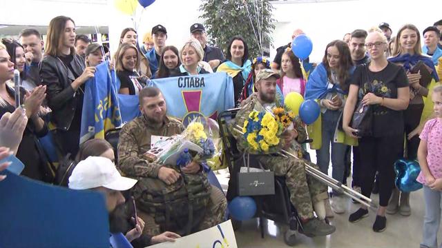 Two Ukrainian men sit in wheelchairs, holding bouquets of flowers, and surrounded by a crowd of people, some wearing Ukrainian flags or holding balloons. 