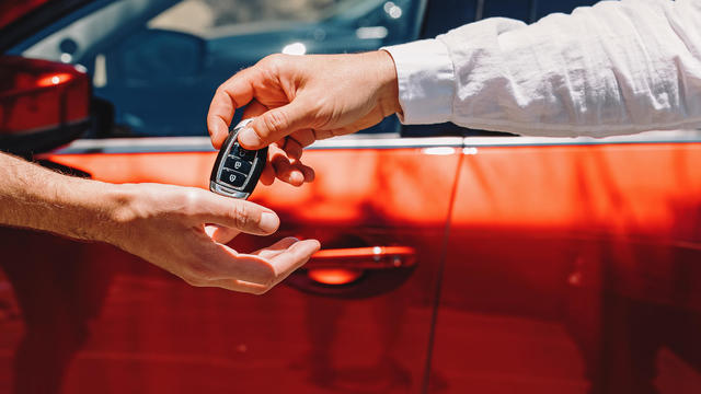 Dealer Giving Car Keys To The New Owner. Close-Up Of Hands On The Background Of Red Automobile 
