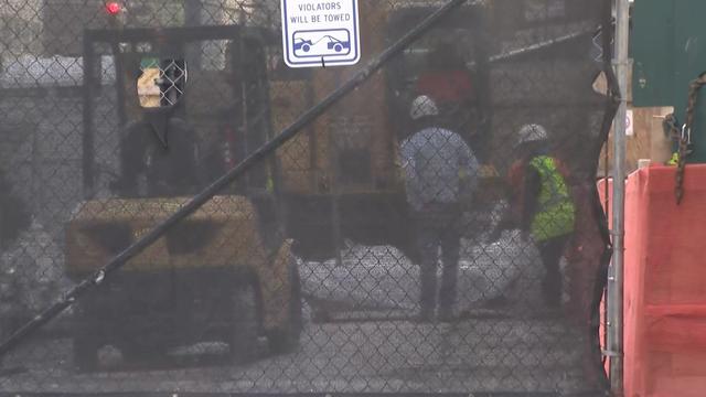 Individuals in hard hats work behind a covered fence. 