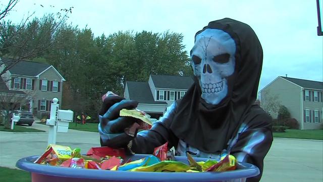 A child wearing a skeleton mask and costume takes candy out of a bucket in a residential cul-de-sac. 