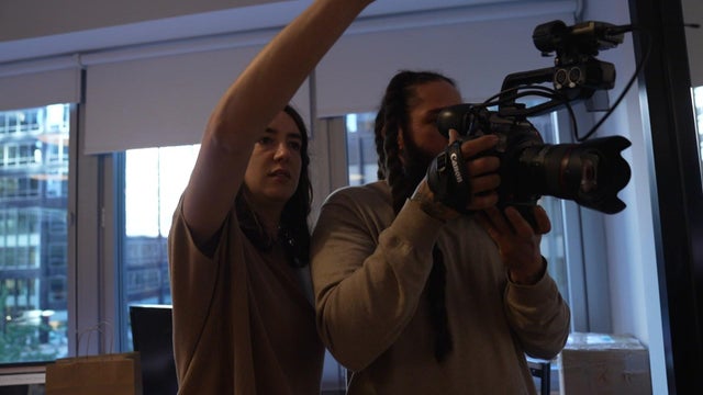 An individual holds a camera while another person offers direction. 