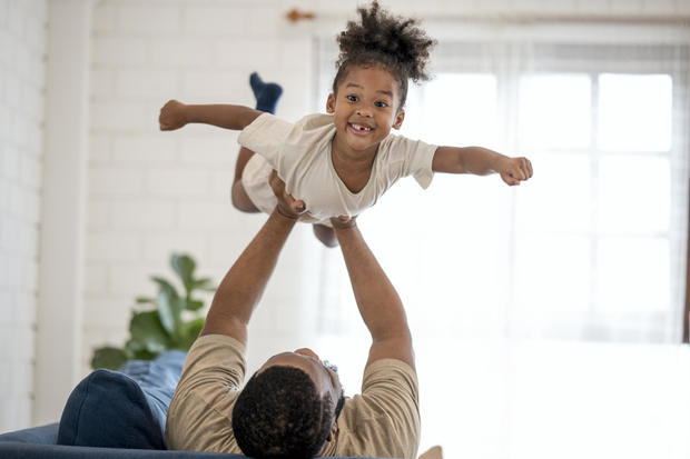 I'm Supergirl!. Happy African cute girl playing with her father on sofa at home. Family happily spend time together. 