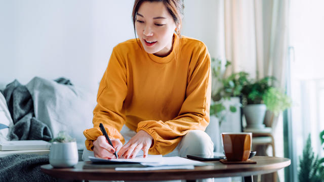 Young Asian woman holding a pen and signing paperwork in the living room at home. Deal concept 