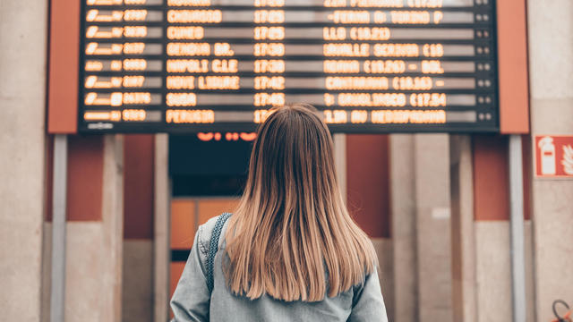 A young woman at a railway station or at the airport looks at the smartphone screen against the background of the arrival and departure board 