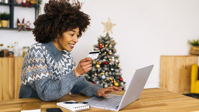 Happy young woman with credit card using laptop sitting at table in living room 