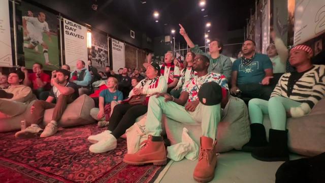 Dozens of people sit in bean bag chairs and on the floor, watching a large screen playing a World Cup game. 