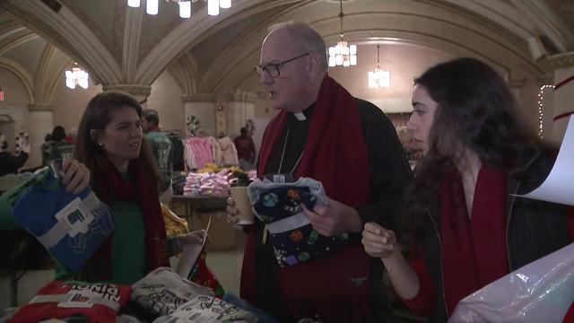 Timothy Cardinal Dolan and two volunteers stand at a table of clothes. 