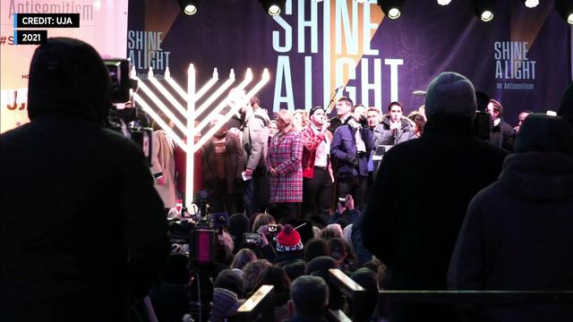 Spectators stand in front of a crowded stage and a brightly lit menorah. Signage says "Shine a light on antisemitism." 