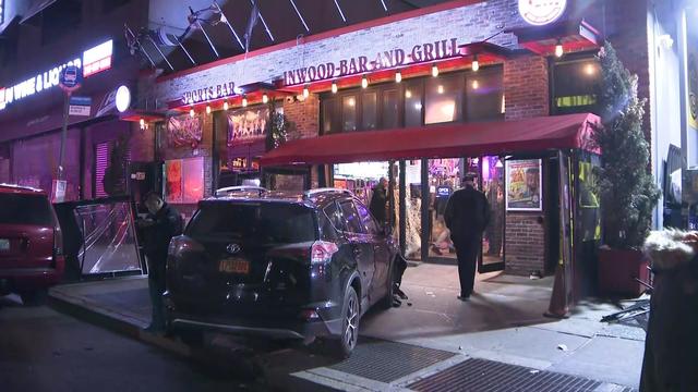 A vehicle with front-end damage sits on a sidewalk in front of a restaurant with a shattered window. 