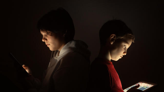 Brother and sister use smartphone and digital tablet while sitting back to back at night at home 