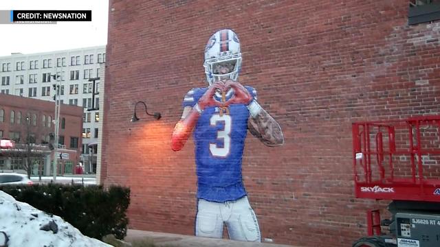 A mural of Buffalo Bills safety Damar Hamlin in his uniform making a heart shape with his hands is painted on the side of a red brick wall. 