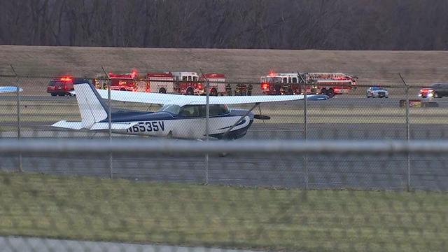 A small plane can be seen parked at an airport. Multiple fire trucks and police vehicles can be seen in the background. 