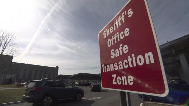 Vehicles drive past a sign reading "Sheriff's Office Safe Transaction Zone." 