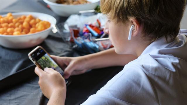 Preteen boy concentrating on smartphone with ear buds and bowls of snacks 