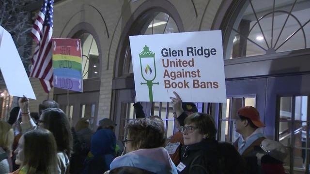 People stand outside a building holding signs, including one reading, "Glen Ridge United Against Book Bans." 