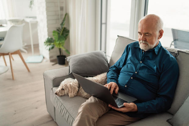 Senior man sitting on sofa with his dog and using laptop at home. 