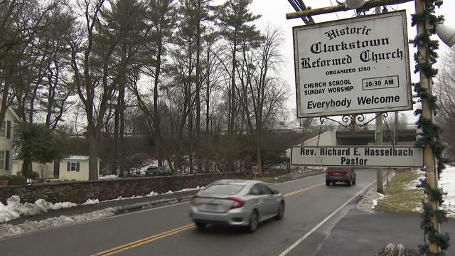 A sign for the Historic Clarkstown Reformed Church is in the foreground along a residential road. In the background, the New York State Thruway can be seen. 