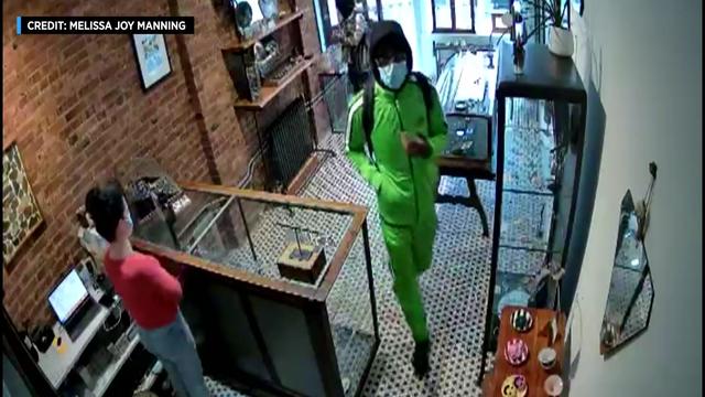 A man in a bright green track suit, face mask and sunglasses walks behind the counter at a Brooklyn jewelry store. 