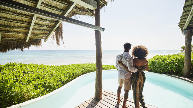 Wide shot rear view of couple standing poolside at luxury tropical beachfront villa looking at view 